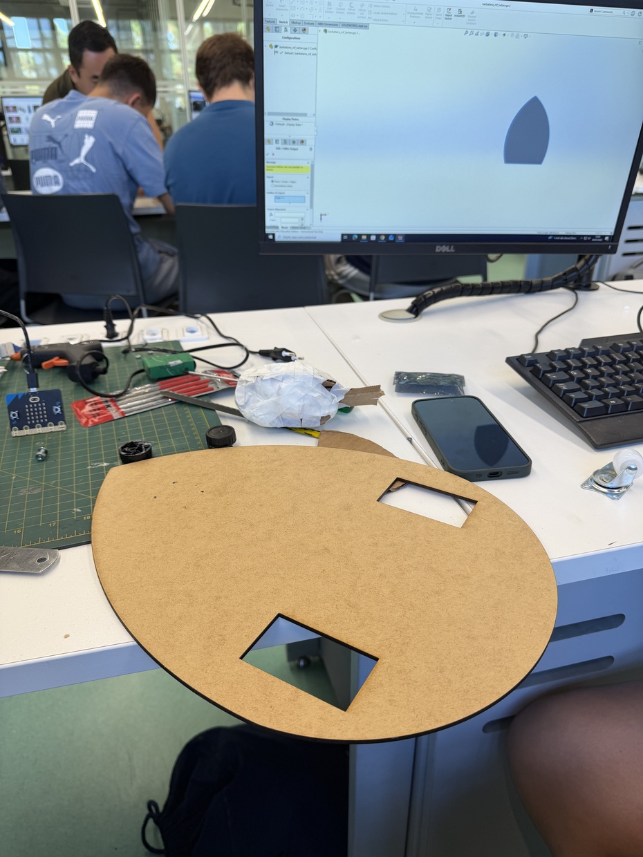 Montagem Mecânica (base):

A base da tartaruga (chassi) foi realizada no software SolidWorks e cortada utilizando a maquina de corte à laser utilizando uma placa de MDF com formato semelhante a oval, e tamanho de 25x30cm, mas também podendo ser feito com impressão 3D ou papelão.

Durante o projeto da base, foram criadas duas aberturas retangulares (“janelas”) para o encaixe das rodas motorizadas, permitindo que parte das rodas ficasse exposta para contato direto com o solo, reduzindo a altura total do robô e melhorando sua estabilidade. Essas janelas também auxiliaram no alinhamento dos motores e evitaram atrito entre as rodas e a estrutura de MDF. Após o corte da peça, foram realizados furos com furadeira manual para fixação dos componentes mecânicos, principalmente da roda boba frontal, instalada com parafusos para garantir equilíbrio e apoio durante a movimentação.