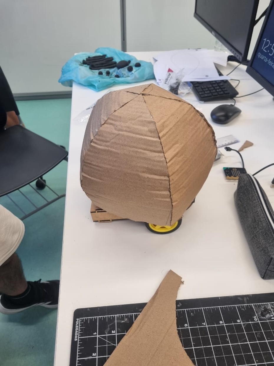 Acesse o site Templatemaker e selecione um molde de semi esfera para servir como base do corpo da aranha.
Defina as dimensões desejadas da semi esfera de acordo com o tamanho do robô.
Gere o molde no site e faça o download do arquivo em PDF.
Imprima o molde em folha sulfite e recorte todas as partes indicadas.
Transfira o molde para o papelão utilizando lápis ou caneta para marcar os contornos.
Recorte cuidadosamente as peças do papelão usando tesoura ou estilete.
Dobre as abas do molde para facilitar a montagem da semi esfera.
Utilize cola quente para unir as partes do papelão até formar a estrutura arredondada do corpo da aranha.
Verifique se a estrutura ficou firme e bem alinhada.
Prepare o papel machê utilizando:
tiras de jornal,
cola branca,
água.
Misture cola branca e água até formar uma solução homogênea.
Mergulhe as tiras de jornal na mistura e aplique sobre toda a estrutura de papelão.
Faça várias camadas de papel machê para deixar o corpo mais resistente.
Aguarde a secagem completa entre uma camada e outra.
Após secar totalmente, lixe levemente a superfície para deixá-la mais uniforme.
Utilize cola quente para fixar o corpo da aranha na estrutura principal do robô.
Pinte toda a estrutura utilizando tinta acrílica preta e pincel.
Aguarde a secagem da tinta e aplique uma segunda camada, caso necessário.
Faça os acabamentos finais, corrigindo imperfeições e reforçando partes soltas com cola quente.
Finalize verificando se o corpo da aranha está firme, centralizado e bem preso ao robô.